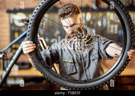 Handsome repairman in workwear serving mountain bicycle, standing with front wheel at the workshop of a bicycle shop