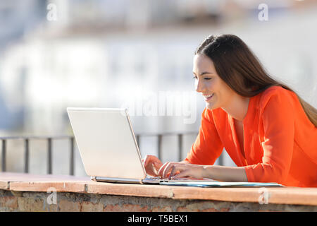 Happy entrepreneur in orange working writing on laptop in a balcony Stock Photo