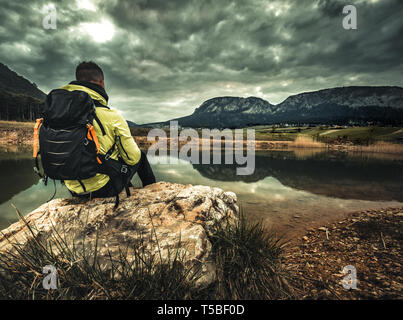 A person sits on the edge of a cliff overlooking a blue alpine lake on a cloudy day Stock Photo