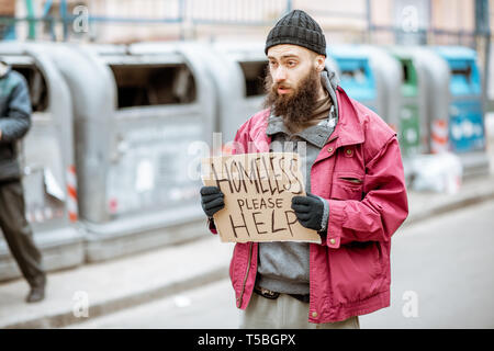 Portrait of a depressed homeless beggar standing on the street with social message on the cardboard. Concept of human poverty Stock Photo