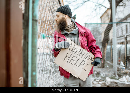 Portrait of a depressed homeless beggar standing with social message on the cardboard near the old fence outdoors Stock Photo