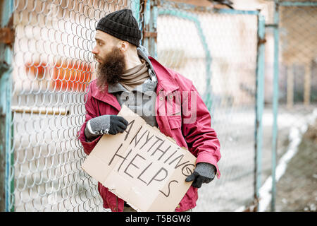 Portrait of a depressed homeless beggar standing with social message on the cardboard near the old fence outdoors Stock Photo