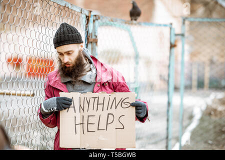 Portrait of a depressed homeless beggar standing with social message on the cardboard near the old fence outdoors Stock Photo