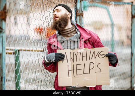 Portrait of a depressed homeless beggar standing with social message on the cardboard near the old fence outdoors Stock Photo