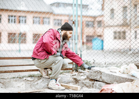 Portrait of a depressed homeless beggar or prisoner standing near the old metal fence outdoors Stock Photo