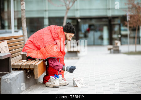 Depressed homeless sitting on the bench wrapped with sleeping bag asking some money near the business center Stock Photo