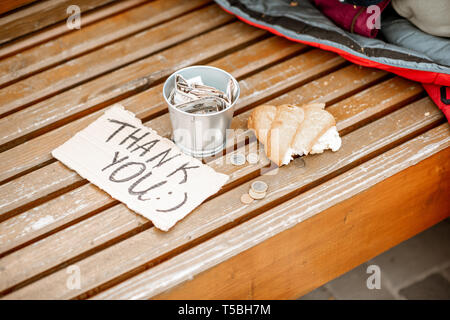 Beggar's cup with thank you message and bread on the bench outdoors. Begging money concept Stock Photo