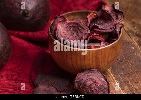 Close-up beetroot chips in wooden bowl and whole beetroots on red napkin on old wooden table. Stock Photo