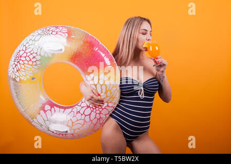 Pretty young blonde in swiming suit enjoying her orange cocktail over yellow background. Tasty cocktail. Swim ring. Fit body . Stock Photo