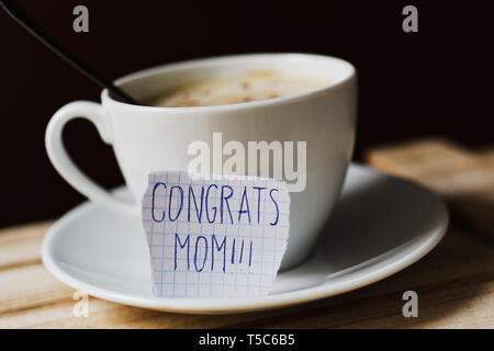 closeup of a piece of paper with the text congrats mom written in it, next to a white ceramic cup with cappuccino Stock Photo