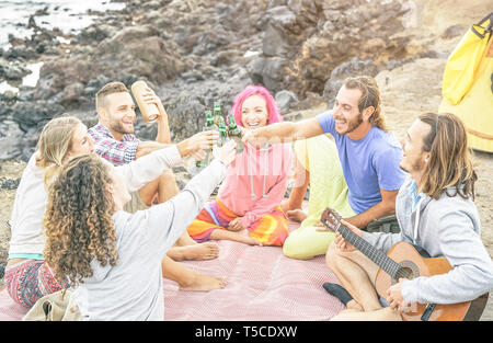 Group of happy friends toasting beers and playing guitar camping with tent on the beach - Travel people having fun listening music and drinking Stock Photo