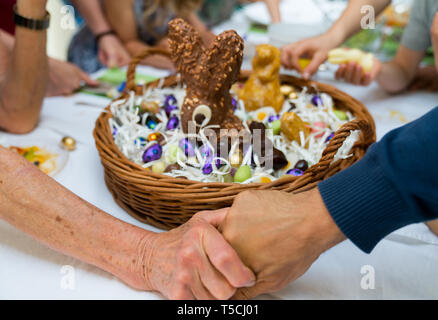 grandmother joins hands with son and gives thanks during Easter with a large Easter egg basket filled with candy and many relatives behind Stock Photo