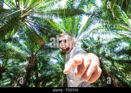 Bearded smiling man with glasses standing in the palm tree forest and pointing his finger to the camera. Stock Photo