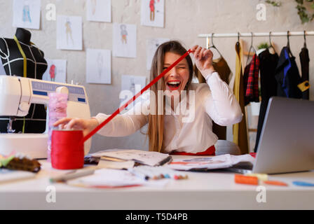 Photo of happy female dressmaker unwinding red ribbon sitting at table in atelier Stock Photo