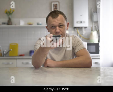 Young guy with a TV remote control switches channels while sitting in the kitchen, front view Stock Photo