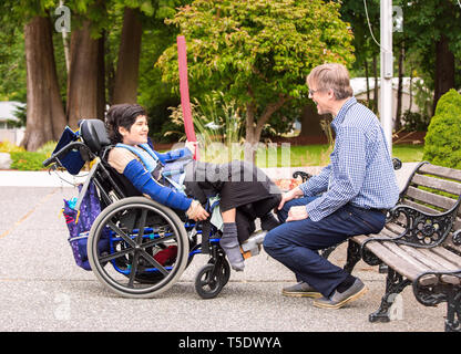 Caucasian father talking and laughing with twelve year old disabled biracial son in wheelchair outdoors on sunny day at park Stock Photo