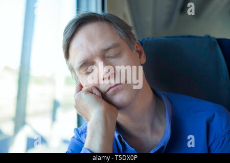Caucasian man wearing blue shirt in early fifties sleeping and resting head on hand near train window Stock Photo