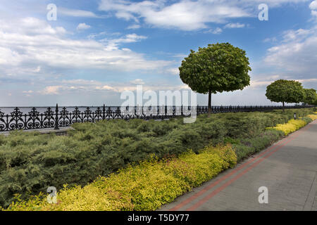 A paved stepped road in a beautiful park passes along the embankment of the river with ornamental flowers, framed decorative fence with cut bushes and Stock Photo