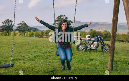 Happy young woman swinging in a recreational area with motorcycle in the background