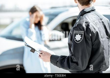 Policeman issuing a fine for violating the traffic rules to a young upset woman driver stadning near the car on the background Stock Photo