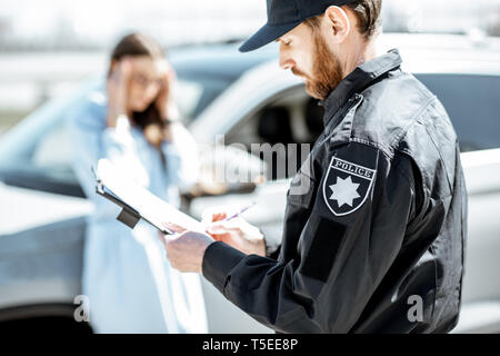 Policeman issuing a fine for violating the traffic rules to a young upset woman driver stadning near the car on the background Stock Photo