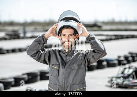 Portrait of a happy male racer in uniform wearing protective helmet while standing on the track with go-karts outdoors Stock Photo