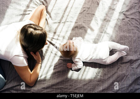 Mother with her newborn son lay on the bed in the rays of sunlight coming out of the window through the curtains. Top view Stock Photo