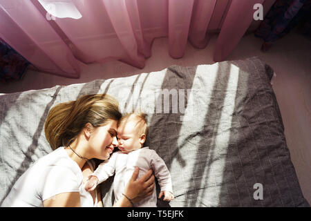 Mother with her newborn son lay on the bed in the rays of sunlight coming out of the window through the pink curtains Stock Photo