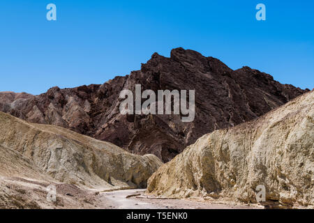 A dry riverbed cuts a pathway through a colorful desert canyon landscape to a rugged mountain peak - Golden Canyon in Death Valley National Park Stock Photo