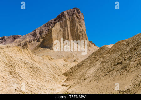 A high desert peak with sheer cliffs rises above a barren, golden desert landscape - Golden Canyon in Death Valley National Park Stock Photo