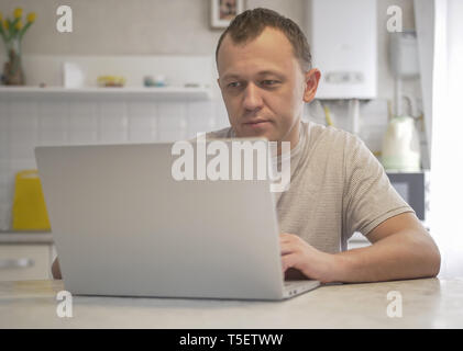 A young man sits in his kitchen with a laptop Stock Photo