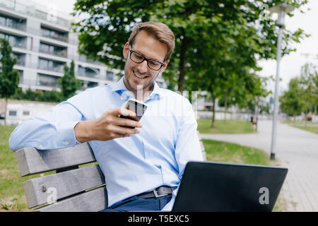 Young businessman sitting on park bench, using smartphone and laptop Stock Photo