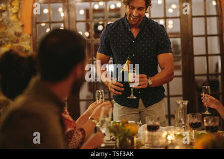 Man serving drinks to friends sitting around table during party. Man pouring champagne into a flute at dinner party. Stock Photo