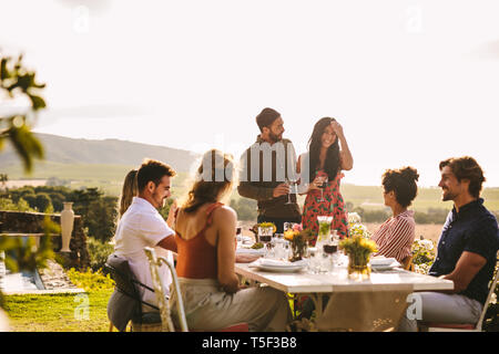 Couple at a party announcing their marriage to friends sitting around dinner table. Smiling woman making an announcement during a dinner party outdoor Stock Photo