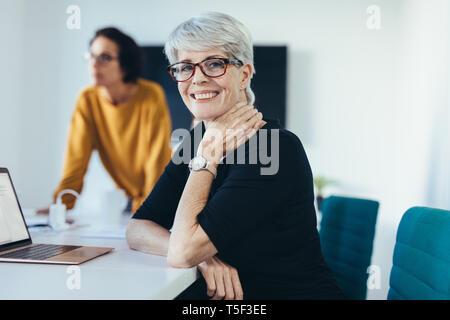 Happy businesswoman sitting at a business meeting with colleagues in background. Woman with coworkers in conference room attending a meeting. Stock Photo