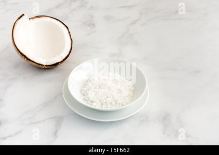 Coconut half and freshly grated coconut flakes in a white porcelain bowl on white marble background with copy space. Stock Photo