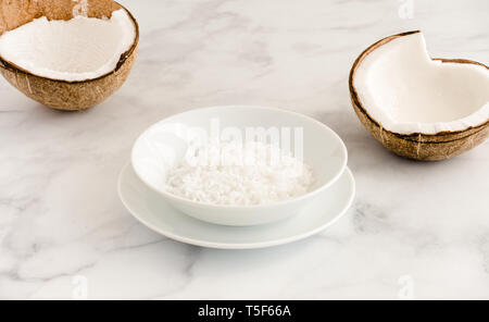 Coconut halves and coconut flakes in a white porcelain bowl on white marble background with copy space. Angled view. Stock Photo