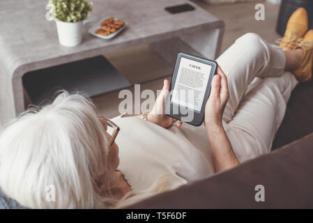 Senior woman sitting by the window, reading an e-book on digital tablet device Stock Photo
