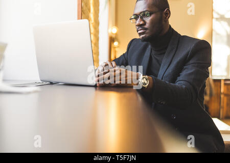 Young handsome dark-skinned businessman in a cafe working behind a laptop with a cup of tea. Stock Photo