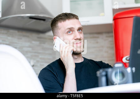 Young and pensive attractive man working on laptop and talking by mobile phone in modern bright apartments. Freelance concept. Home office. Young entr Stock Photo