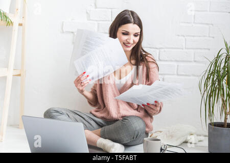 Happy woman examines the paper sitting near the laptop, the girl freelancer works with documents. Student female gets education remotely online Stock Photo
