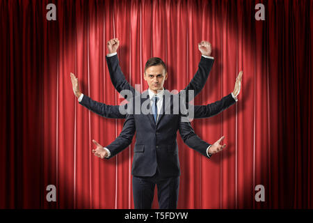Front view of multiarmed businessman raising hands like Shiva, standing lit up by spotlight against red stage curtain. Stock Photo