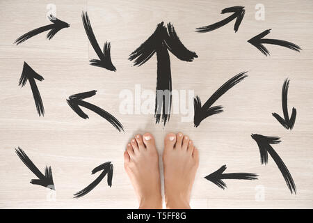 Barefoot woman standing on wooden floor with arrows pointing in different directions. Concept of choice Stock Photo