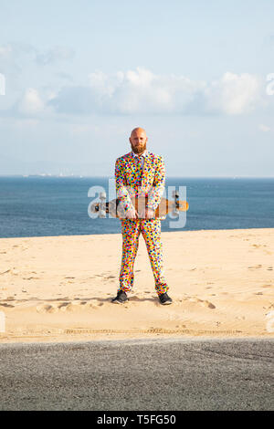 Bearded man with longboard wearing suit with colourful polka-dots standing on sand dune Stock Photo