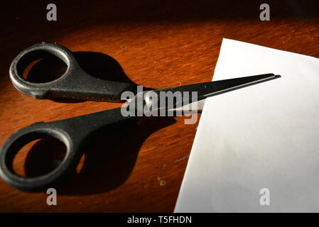 Steel scissors for paper with a piece of white paper, lying on a varnished wooden background. Stock Photo