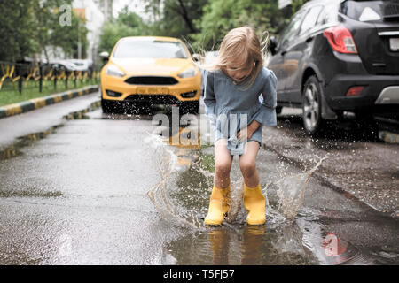 Girl wearing blue dress and rubber boots, jumping in pond on street, yellow car in the background Stock Photo
