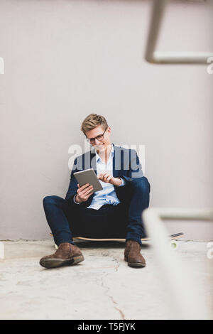 Smiling young businessman with skateboard sitting outdoors using tablet Stock Photo