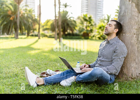 Man with laptop leaning against tree trunk on meadow in city park relaxing Stock Photo