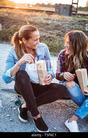 Two laughing friends sitting on longboard eating popcorn Stock Photo
