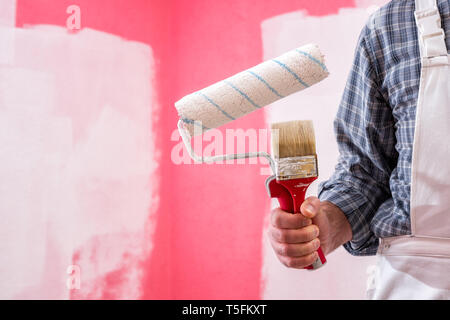 Caucasian house painter worker in white overalls, holding brush and roller for painting. Construction industry. Work tools. Stock Photo